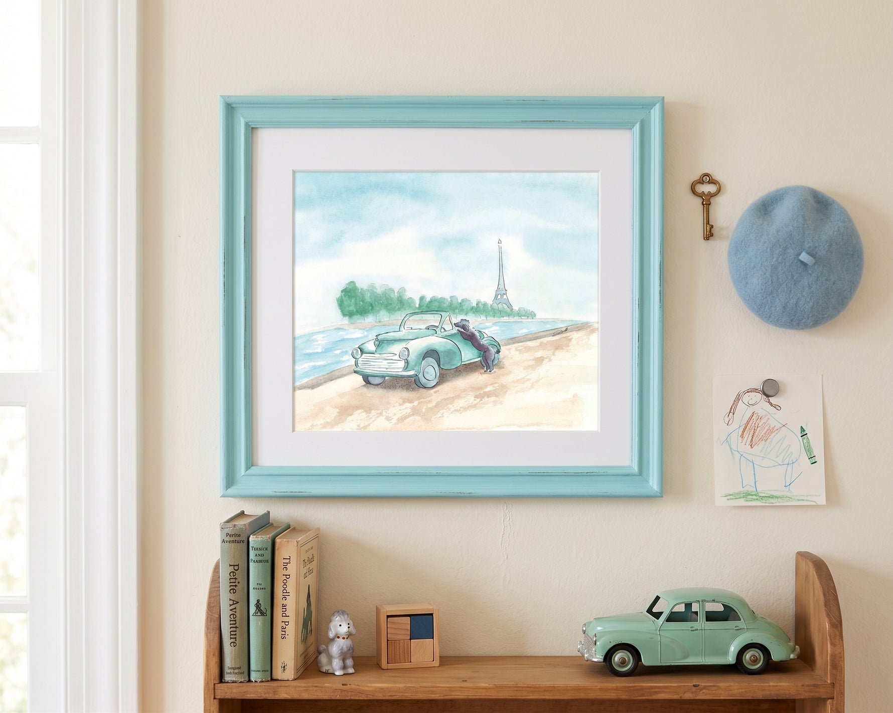 A framed watercolor art print depicting a vintage car on a beach with the Eiffel Tower in the distance, styled on a beige wall above a wooden shelf with books, a toy car, and a blue beret.