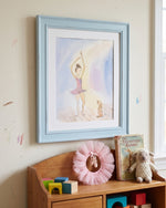 A framed watercolor art print of a ballerina is displayed on a beige wall above a wooden toy shelf with toys and a pink tutu.