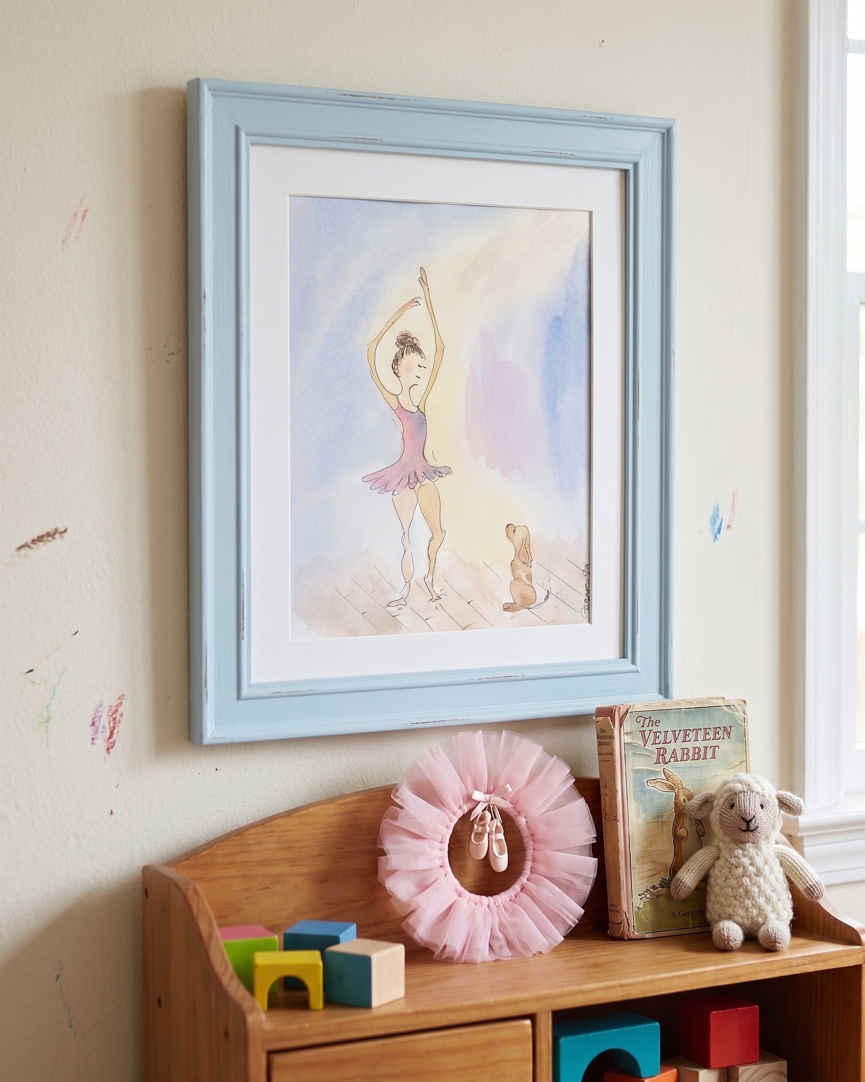 A framed watercolor art print of a ballerina is displayed on a beige wall above a wooden toy shelf with toys and a pink tutu.