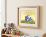 A framed watercolor art print of two purple elephants grazing in a green field, displayed on a cream wall above a wooden shelf with a plush elephant toy, a patterned mug, and colorful blocks.