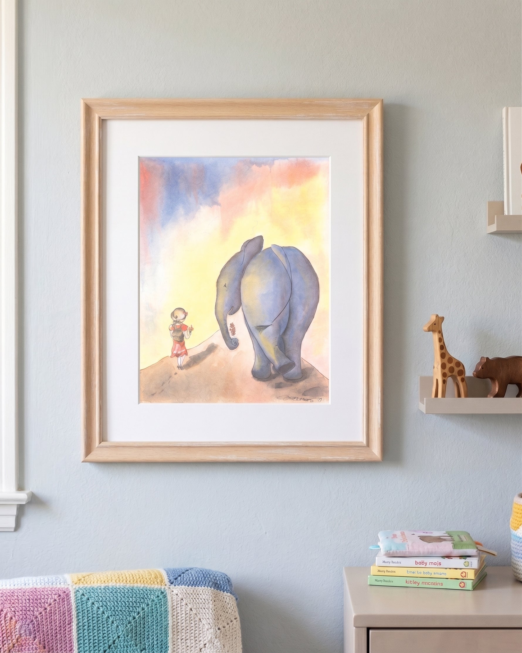A framed watercolor art print depicting a small child and a blue-gray elephant under a pastel sky, displayed on a light gray wall in a styled room setting.
