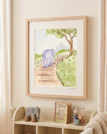 A framed watercolor art print of an elephant on a path, styled on a cream wall above a wooden shelf with toys and a book.