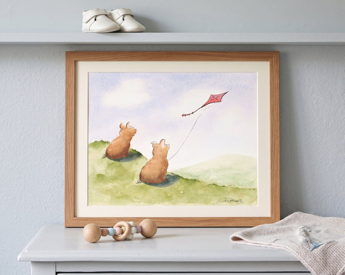 A framed watercolor painting of two teddy bears flying a red kite on a green hill, displayed on a white shelf with baby shoes and a wooden toy.