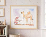 A framed watercolor art print hanging on a light pink star-patterned wallpaper wall. The print features a whimsical illustration of a camel standing next to a woman sewing at a table.