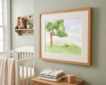A framed watercolor art print of a tree in a grassy field, styled in a nursery setting on a sage green wall above a wooden crib and bedside table.