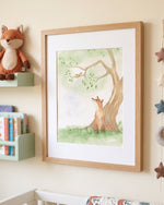 A framed watercolor art print of a fox sitting under a tree, displayed on a nursery wall next to a plush fox toy and a small bookshelf.