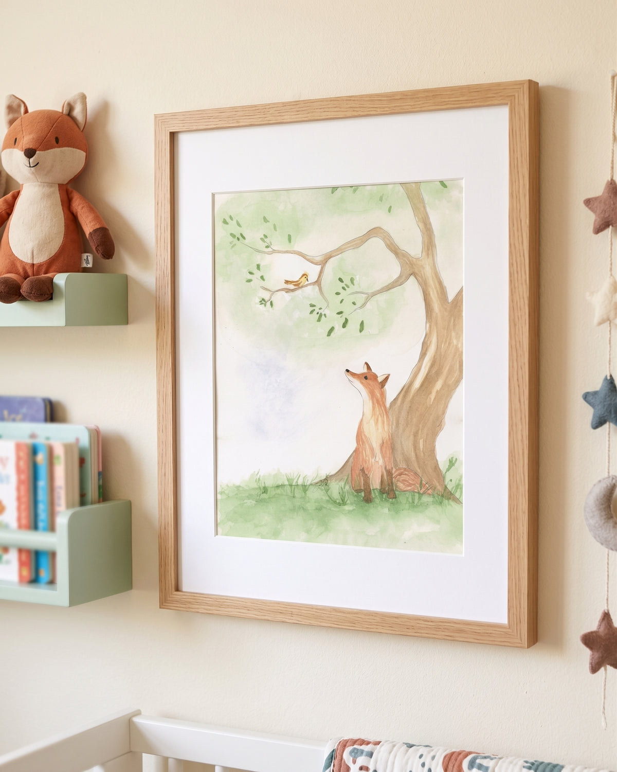 A framed watercolor art print of a fox sitting under a tree, displayed on a nursery wall next to a plush fox toy and a small bookshelf.
