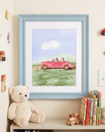 A framed watercolor art print depicting a pink convertible car with two dogs driving on a grassy field under a blue sky, styled on a shelf next to a teddy bear and books.