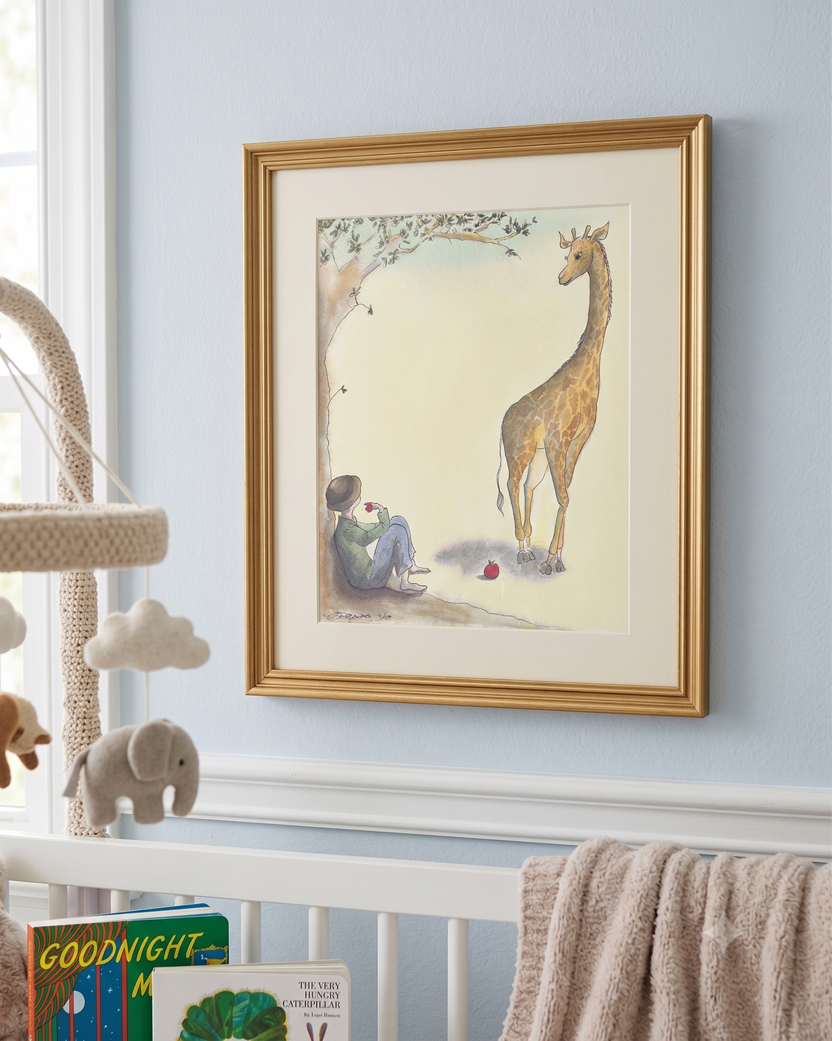 A framed art print featuring a gentle illustration of a child sitting under a tree and a giraffe standing nearby, displayed on a light blue wall above a white crib in a nursery setting.