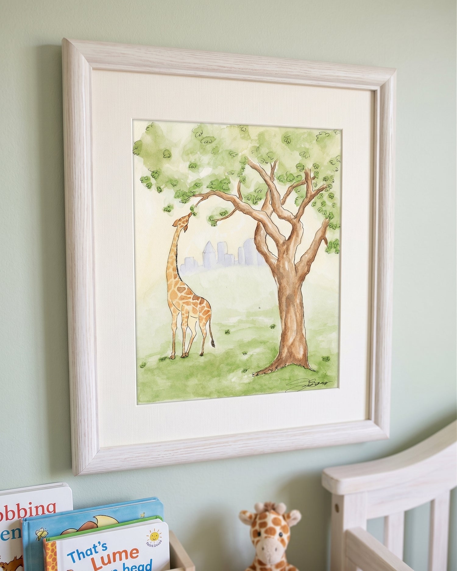 A framed watercolor art print depicting a giraffe and tree, displayed on a light green wall above a crib with baby books and a stuffed giraffe.