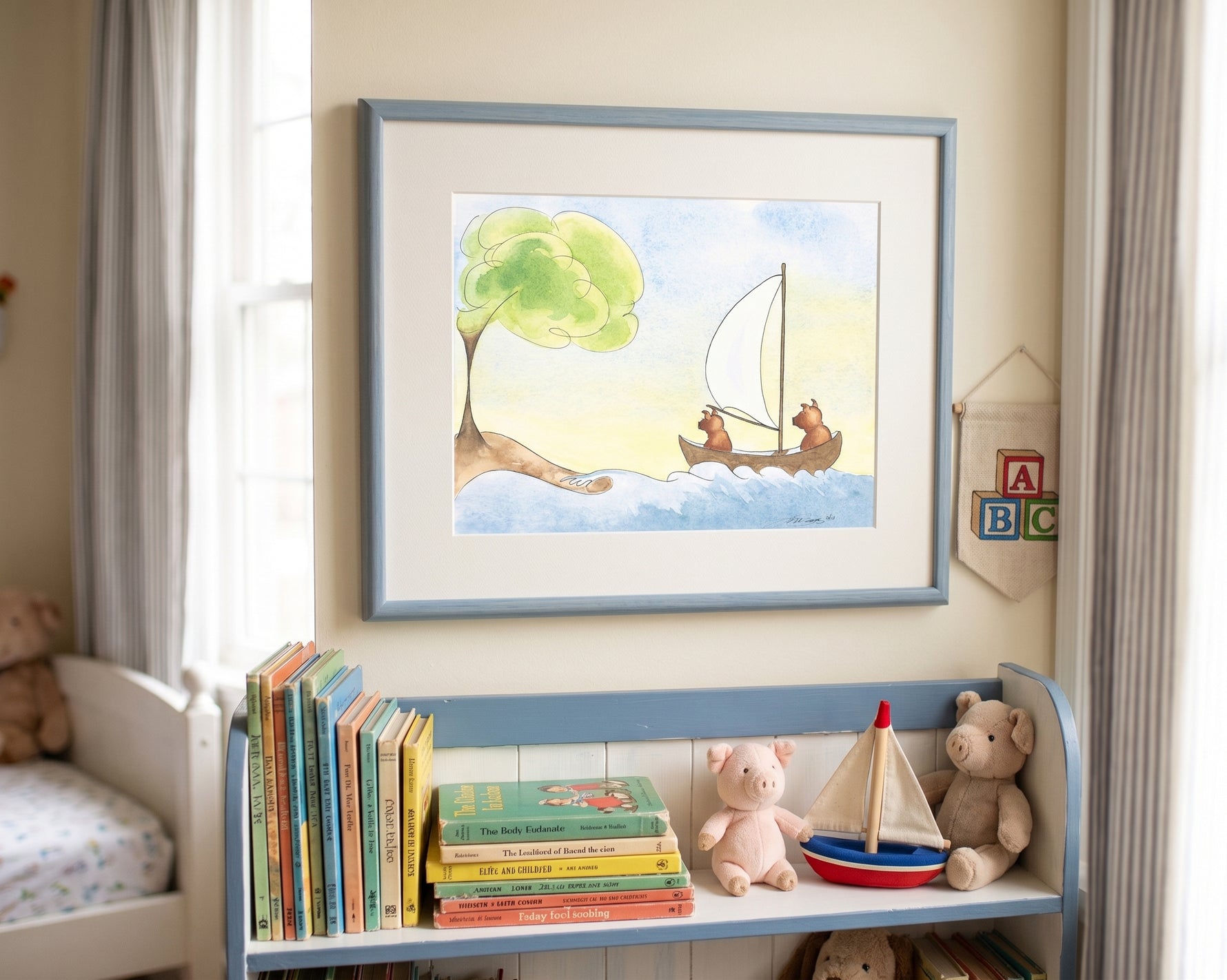 A framed watercolor illustration of two small brown pigs in a wooden sailboat, mounted on a cream-colored mat in a soft blue frame. The picture hangs on a neutral beige wall above a low wooden shelf filled with colorful children's storybooks, a toy wooden sailboat, and plush teddy bears. Natural sunlight filters in from a nearby window draped with light grey curtains.