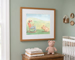 A framed watercolor art print depicting a mother rocking horse and her pig, styled in a nursery setting on a sage green wall above a wooden chest with folded blankets and a teddy bear.